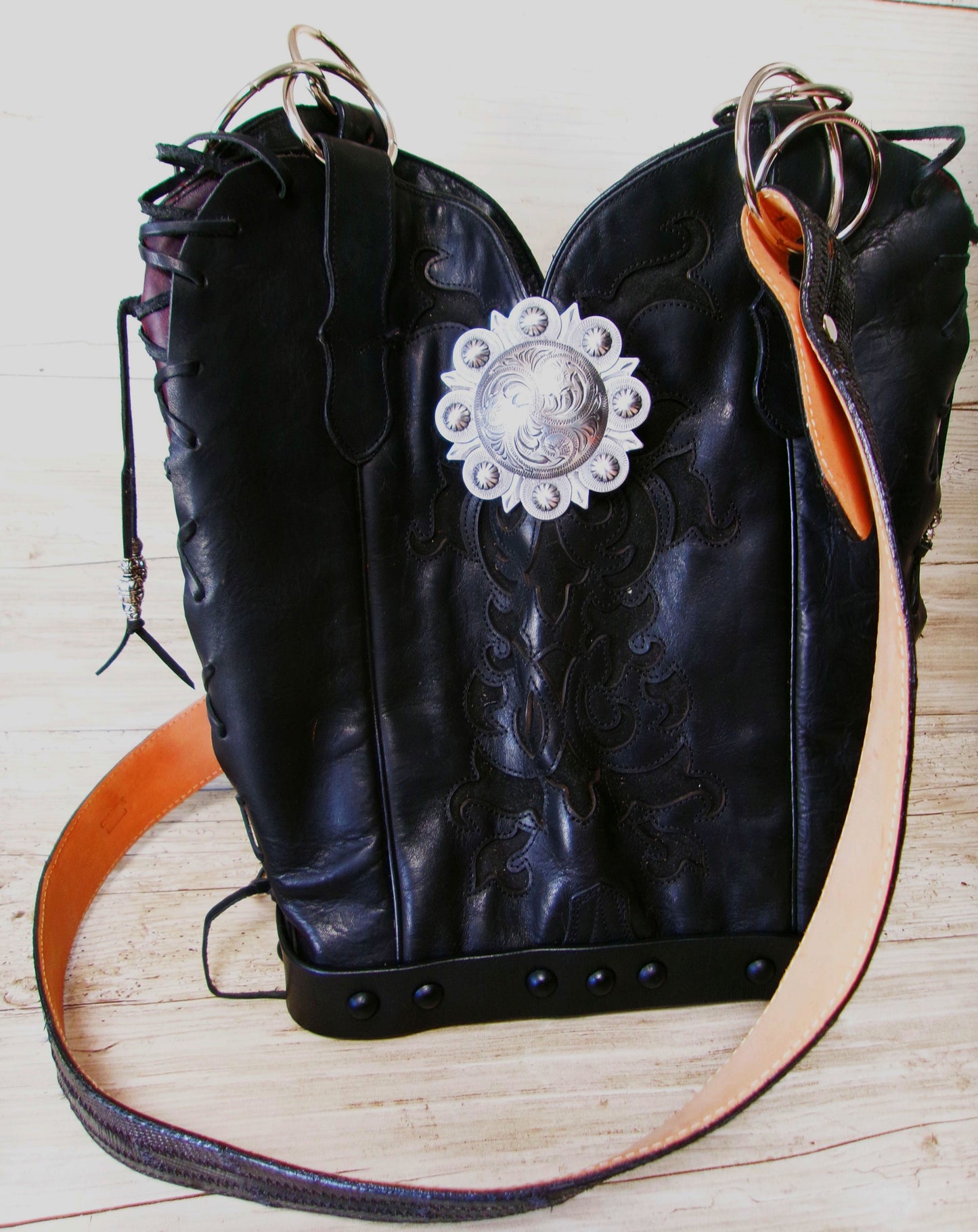 Black leather boot with decorative silver buckle and orange strap on a wooden surface