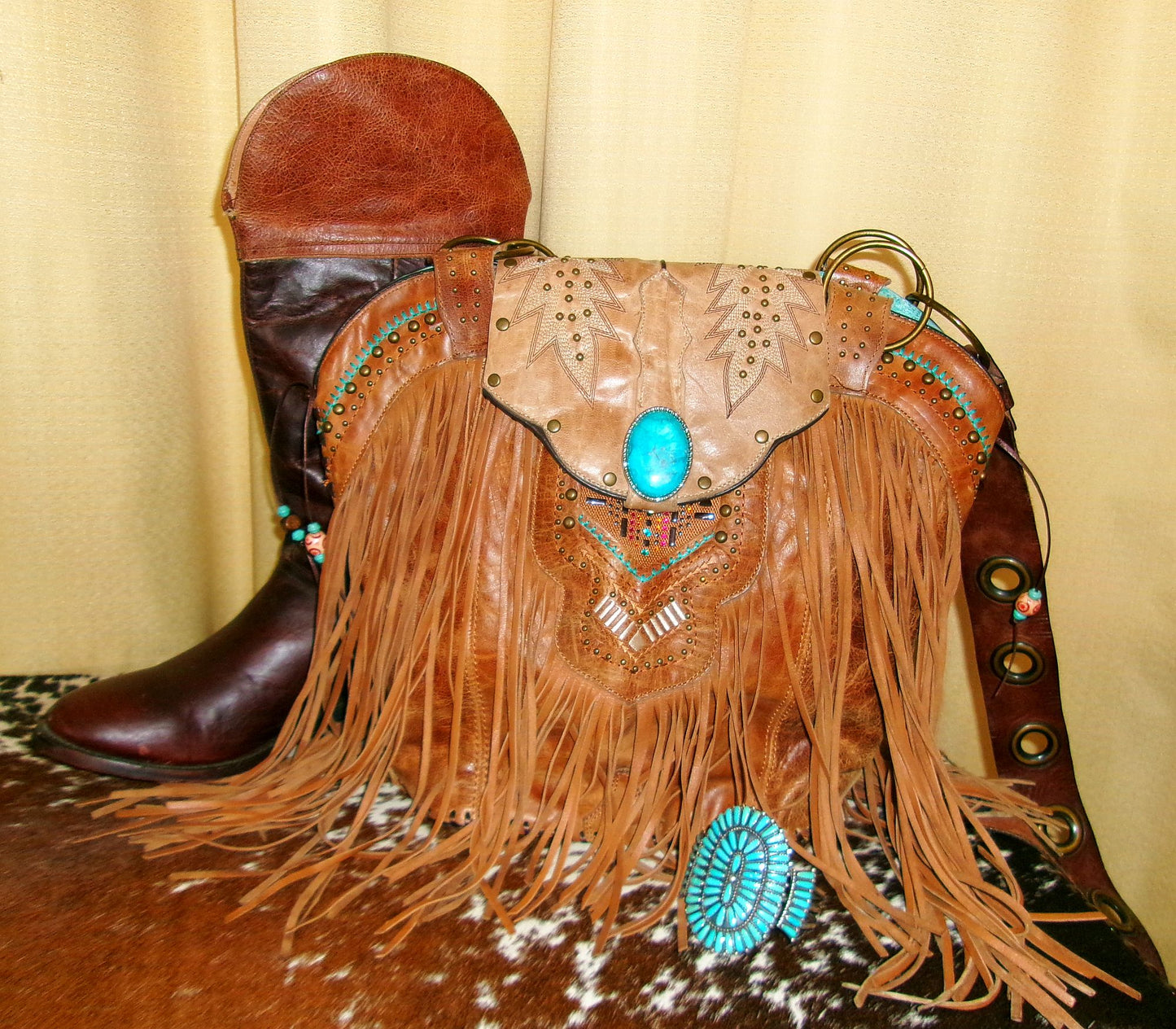 Cowboy boot and decorative leather bag with fringe and turquoise stones on a textured surface.