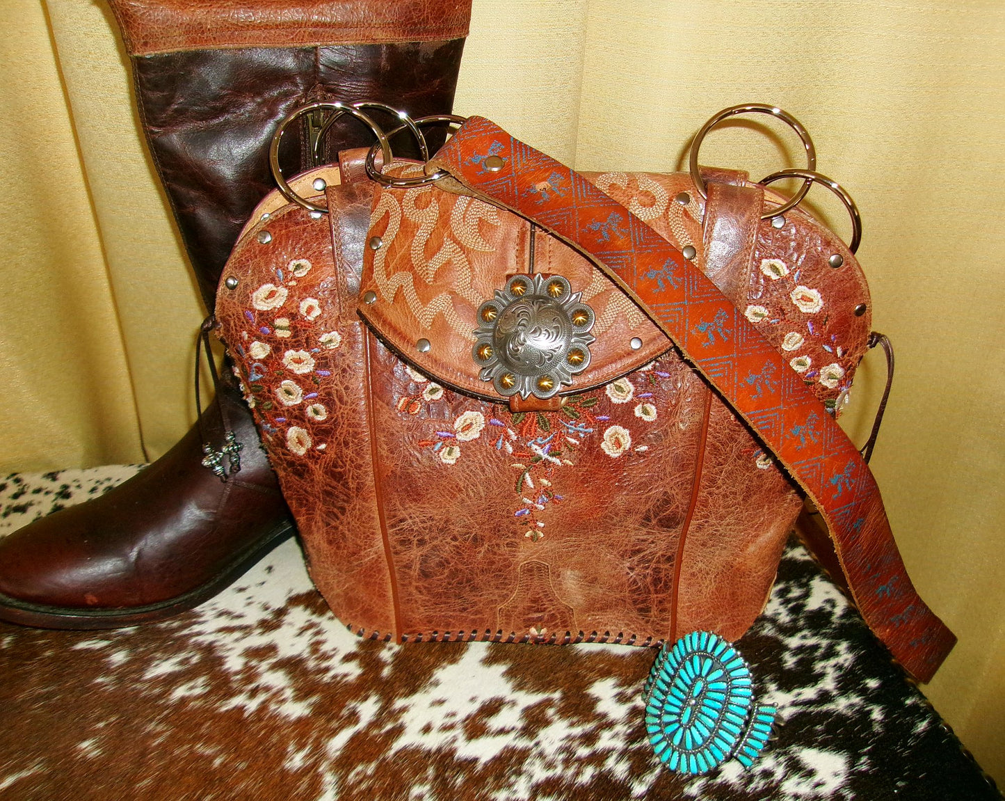 Brown leather bag with floral patterns and a decorative clasp next to a brown boot on a cowhide surface.