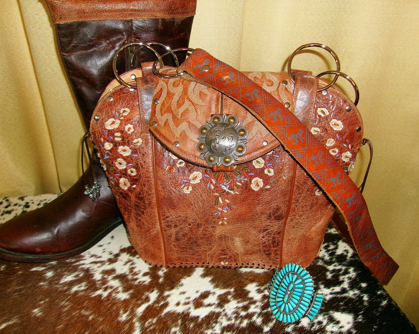 Brown leather bag with floral patterns and a decorative clasp next to a brown boot on a cowhide surface.