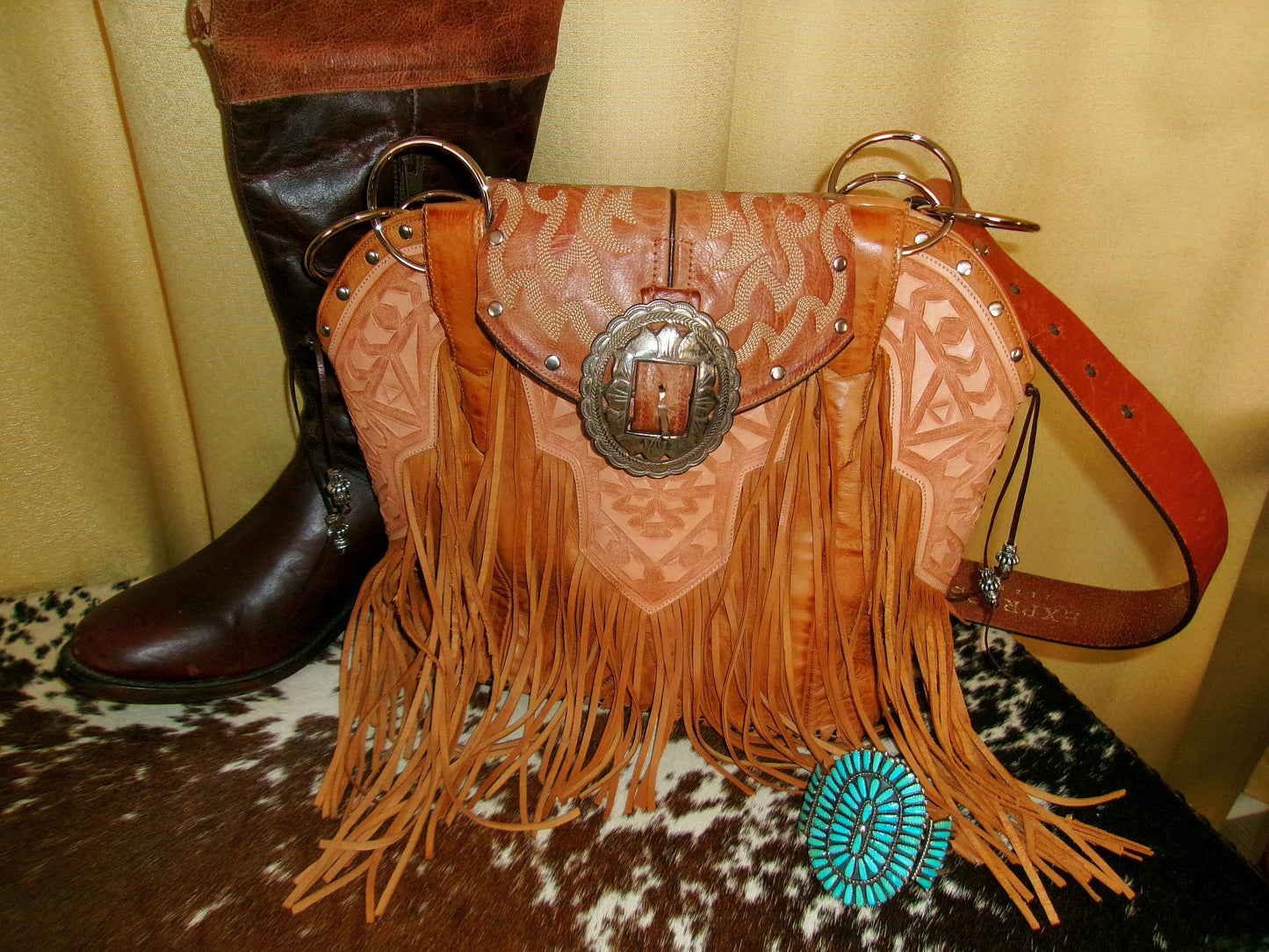 Brown leather handbag with fringe and decorative buckle next to a brown boot on a patterned surface.