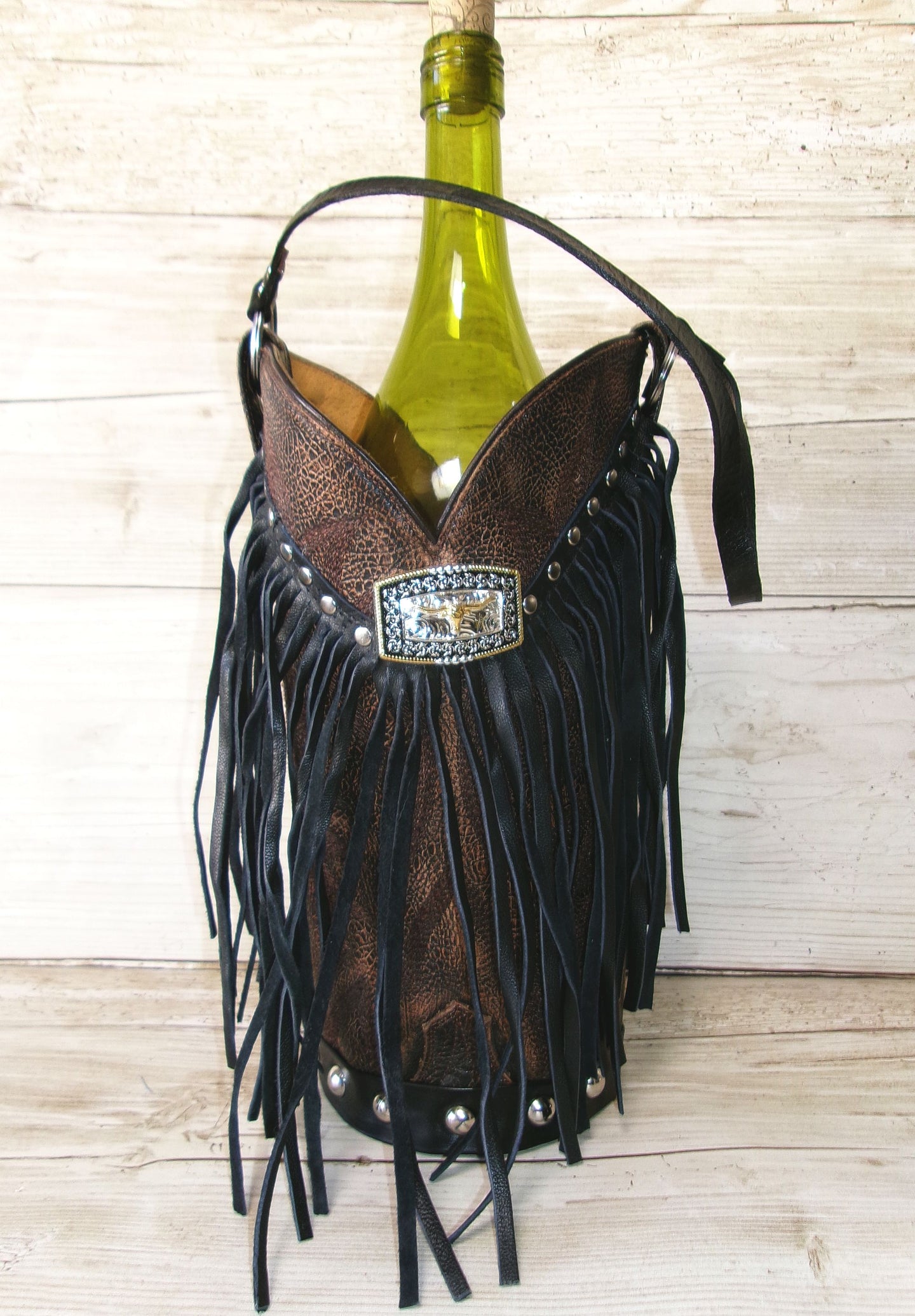 Brown leather wine bottle holder with black fringe and decorative buckle on a wooden surface.